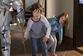 Foto aus dem Film Max und die wilde Sieben: Ein Junge und ein Mädchen schleichen vorsichtig um eine Ritterrüstung herum, die als Dekoration in einem großen Raum steht. 