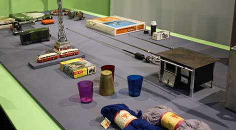 Tisch-ähnlicher Sockel mit Objekten aus verschiedenen Gebieten: Wollknäuel, Becher, ein Spielzeugbus mit Oberleitung ein Kinderherd und Autoantenennen.