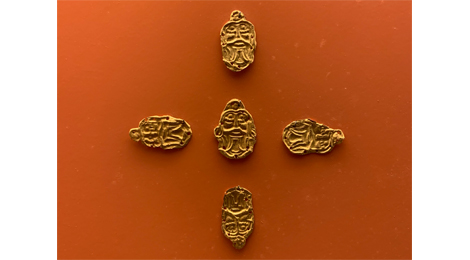 Fünf goldene Plättchen in Form eines Gesichts liegen kreuzförmig auf dem Hintergrund.