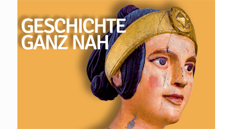 Fahnenmotiv: Ausschnit mit bemaltem Frauenkopf aus Holz und der Beschriftung "Geschichte ganz nah"
