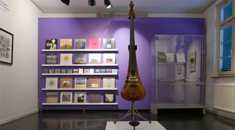 Raum in der Ausstellung "Eberhard Weber. Colours of Jazz" im Stadtmuseum im Gelben Haus. In der Mitte des Raumes steht der Elektrobass von Eberhard Weber, an der Wand stehen in einem Regal Plattenhüllen.