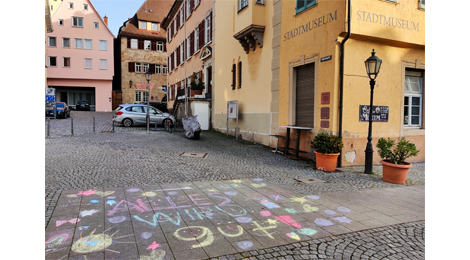Vor dem Stadtmuseum im Gelben Haus ist auf dem Boden eine Zeichnung mit Straßenkreide in verschiedenen Farben. In der Mitte steht „Alles wird gut“. In die Buchstaben sind Gesichter eingezeichnet. Die Worte sind umgeben von kleinen Bildern wie Sonnen, Herzen, Eistüten, Sternen und Punkten.