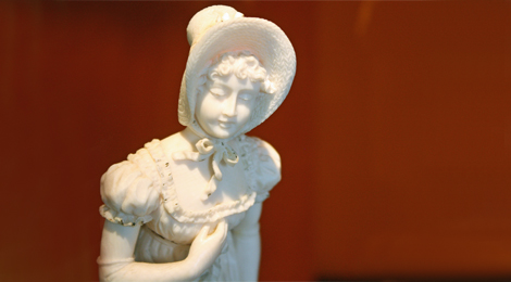 Einfarbige geschnitzte Elfenbeinfigur: Ein Mädchen in einem kurzärmligen Sommerkleid mit Rüschen. Sie hat lockiges Haar und trägt einen Schutenhut mit breiter Krempe.