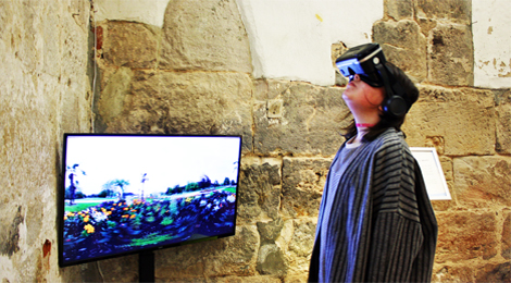 Eine Besucherin trägt einen VR-Brille, die einen Film direkt vor ihre Augen projiziert. Im Hintergrund zeigt ein Monitor eine Gartenlandschaft.